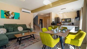 Una sala de estar con un sofá verde, una mesa de centro de madera y sillas de color amarillo brillante. Se ve una cocina al fondo, con un microondas y un televisor.