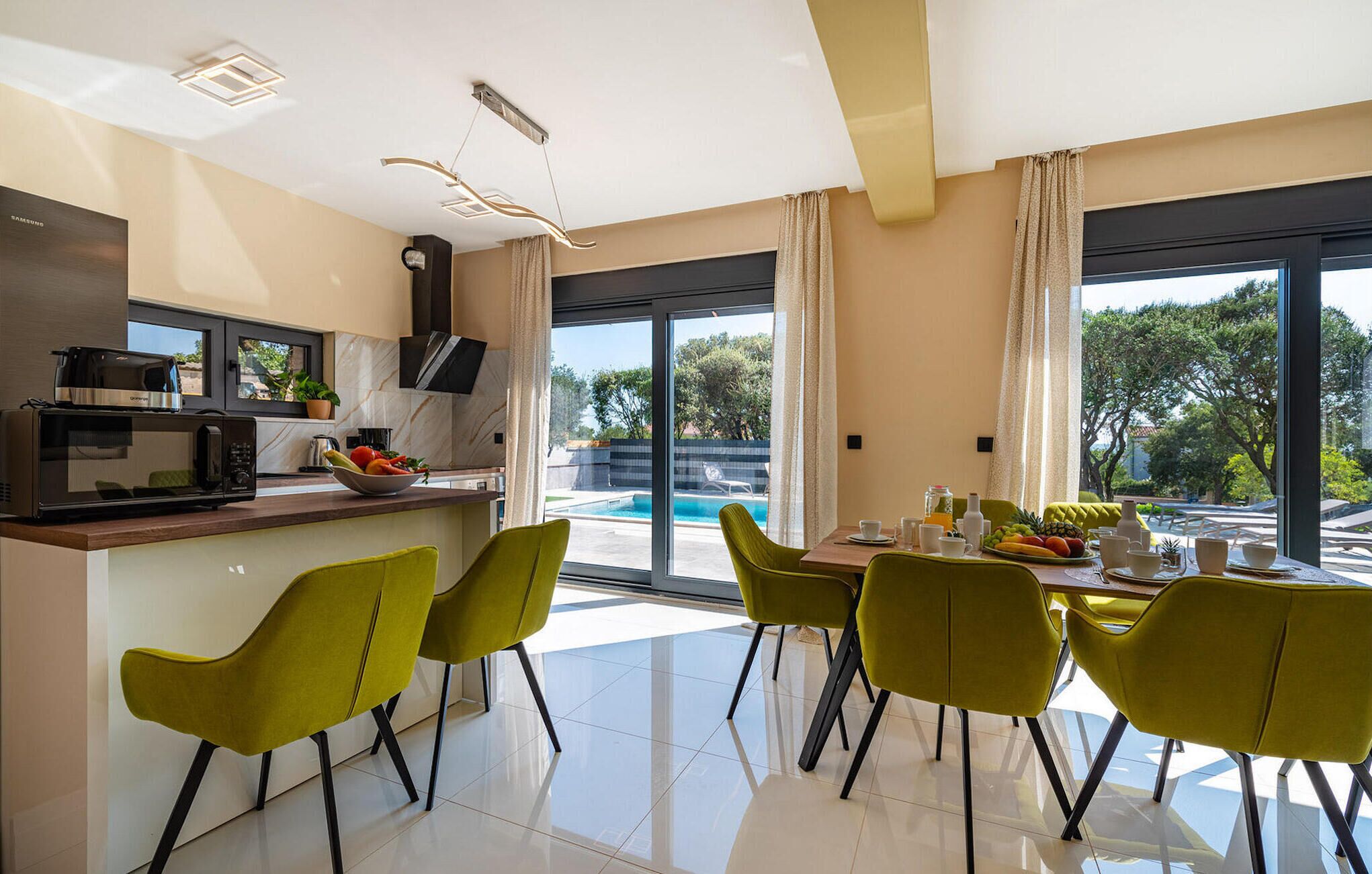Una cocina moderna con microondas integrado, un amplio ventanal con vistas a una piscina y un comedor con sillas verdes y una mesa para el desayuno.