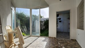 Une pièce dotée d’un sol dallé, d’une porte-fenêtre coulissante donnant sur un jardin et d’un fauteuil placé près de la porte.