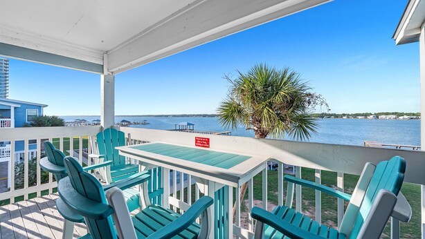 A covered outdoor seating area with chairs and a table, overlooking a body of water with a view of a distant shoreline and buildings. - Family waterfront condo,Dog Friendly, pools, boat launch, beach access, WIFI! (Gulf Shores)