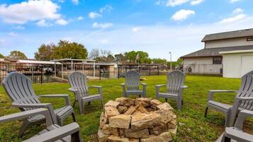 A grassy outdoor area with Adirondack chairs arranged around a stone fire pit.