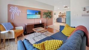Uma sala de estar moderna com uma televisão de tela plana grande, um sofá com almofadas coloridas, uma mesinha lateral e um vaso com planta.