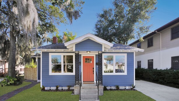 Uma casa pequena, térrea, com porta vermelha, detalhes brancos e fachada azul.