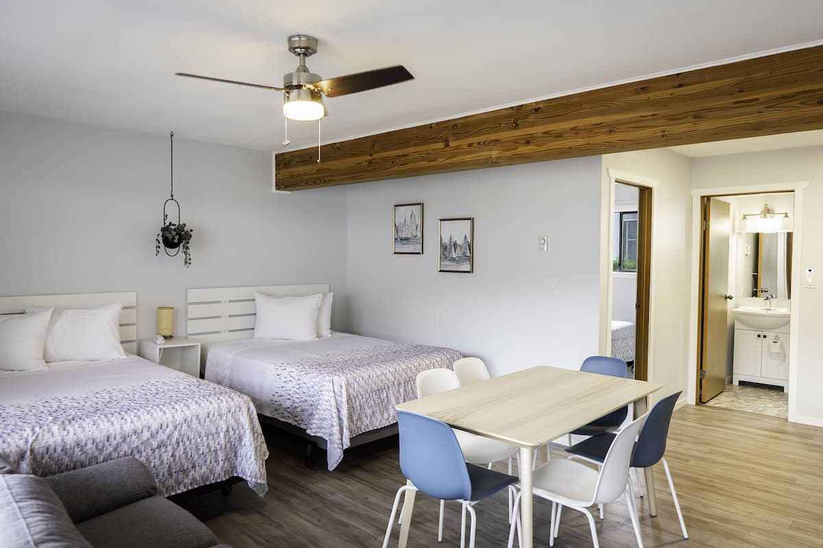 Una habitación de hotel con dos camas, techo de madera, mesa de comedor con sillas, ventilador de techo y una pequeña cocina.