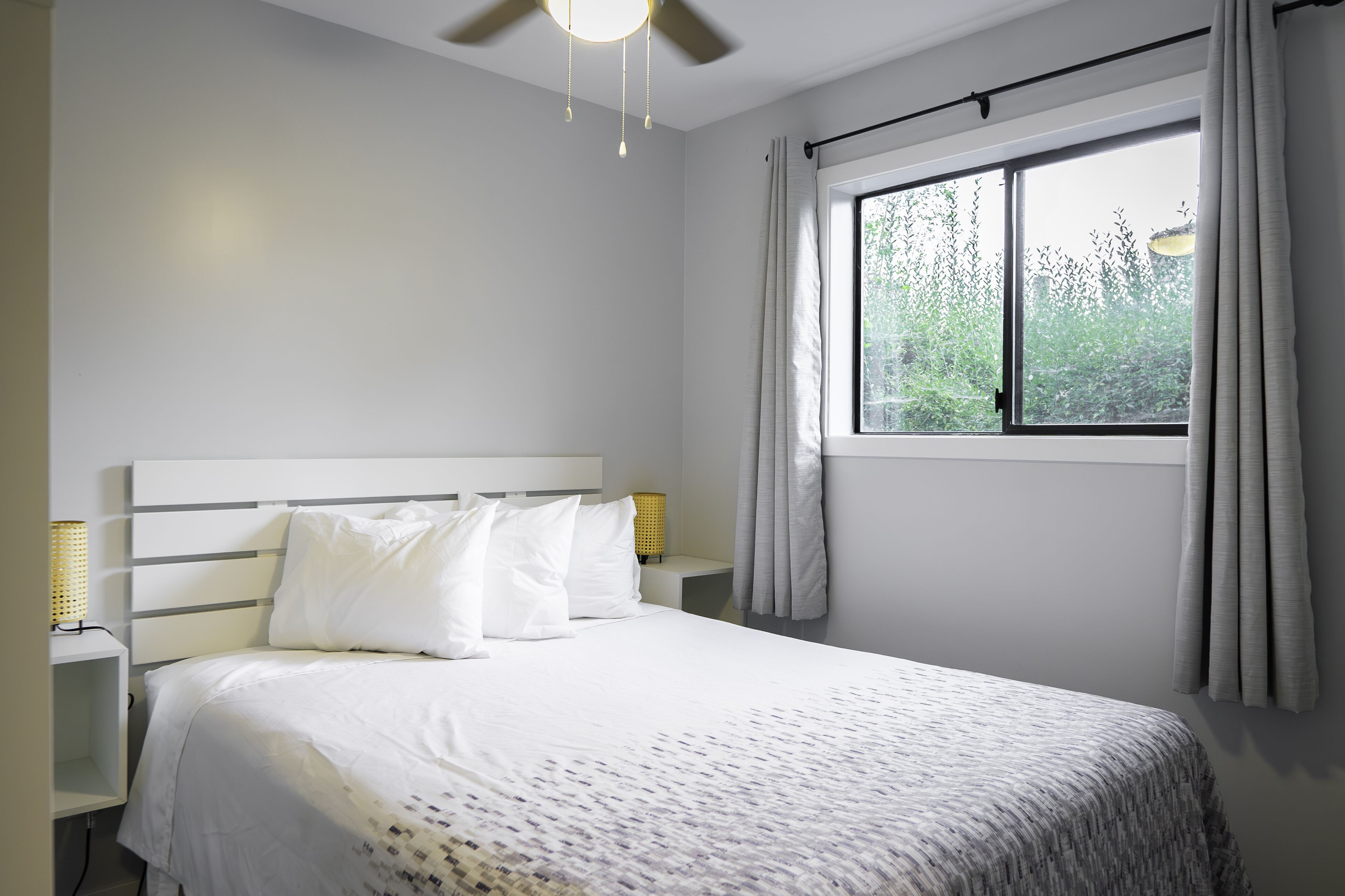 Un dormitorio con cama, mesitas de noche, ventilador de techo y ventana con cortinas.