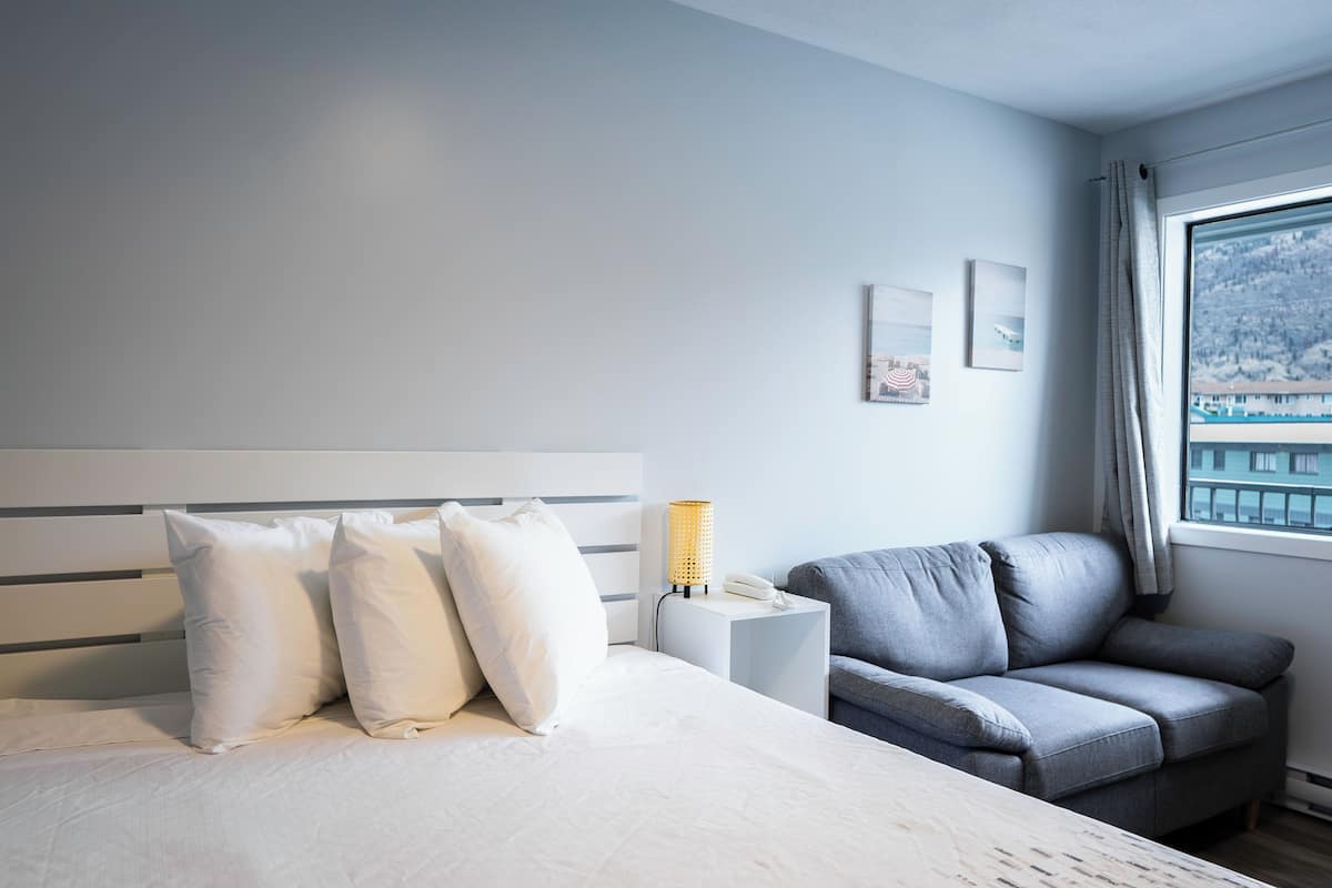 Habitación de hotel con una cama, un sofá gris, una mesita de noche con lámpara y una ventana con vista a un edificio.