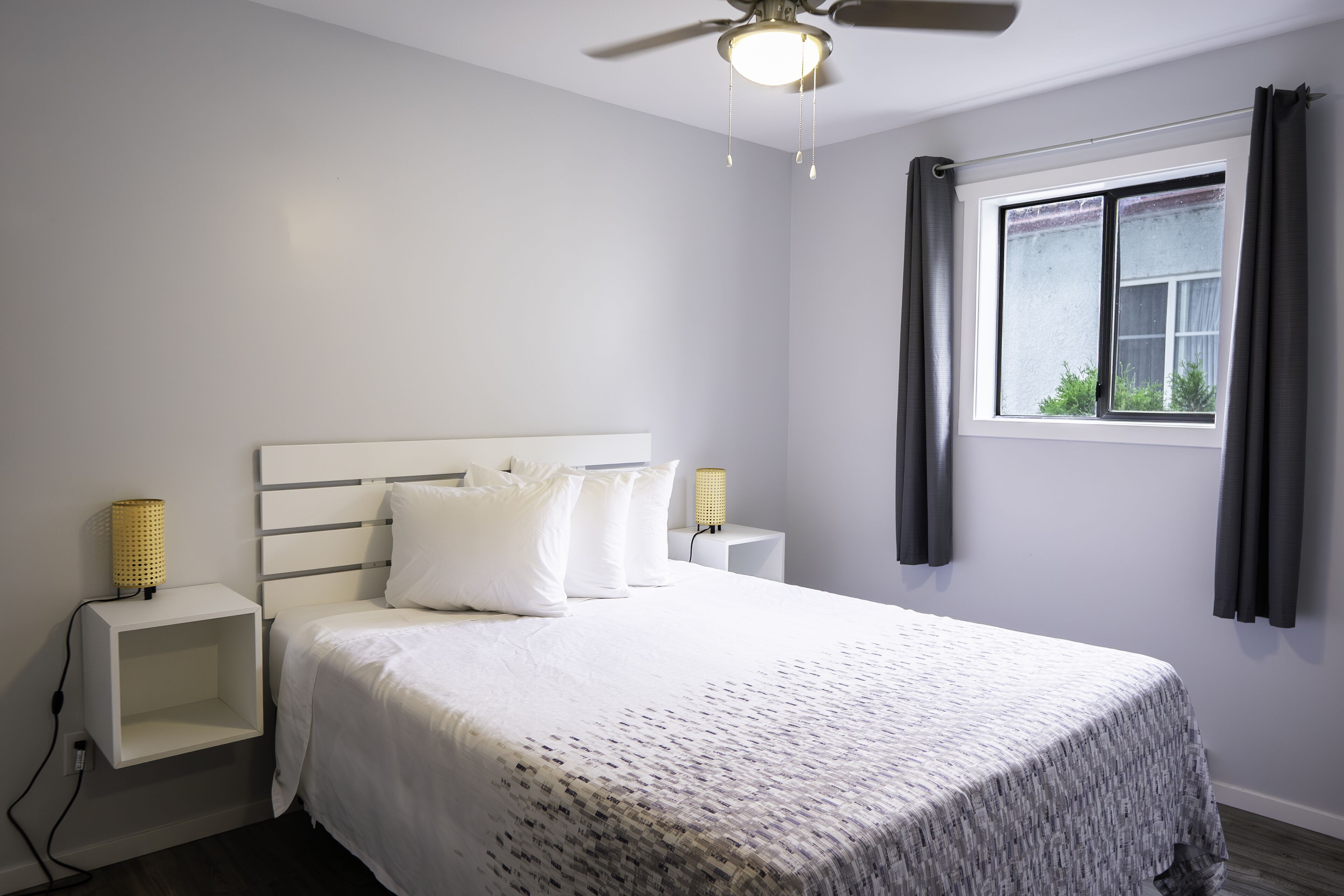 Un dormitorio ordenado con cama, lámparas de noche, ventilador de techo y ventana con cortinas.
