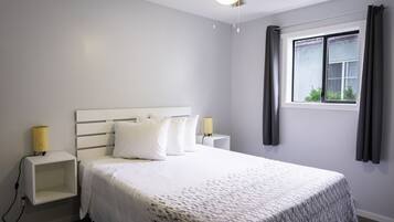 Un dormitorio ordenado con cama, lámparas de noche, ventilador de techo y ventana con cortinas.