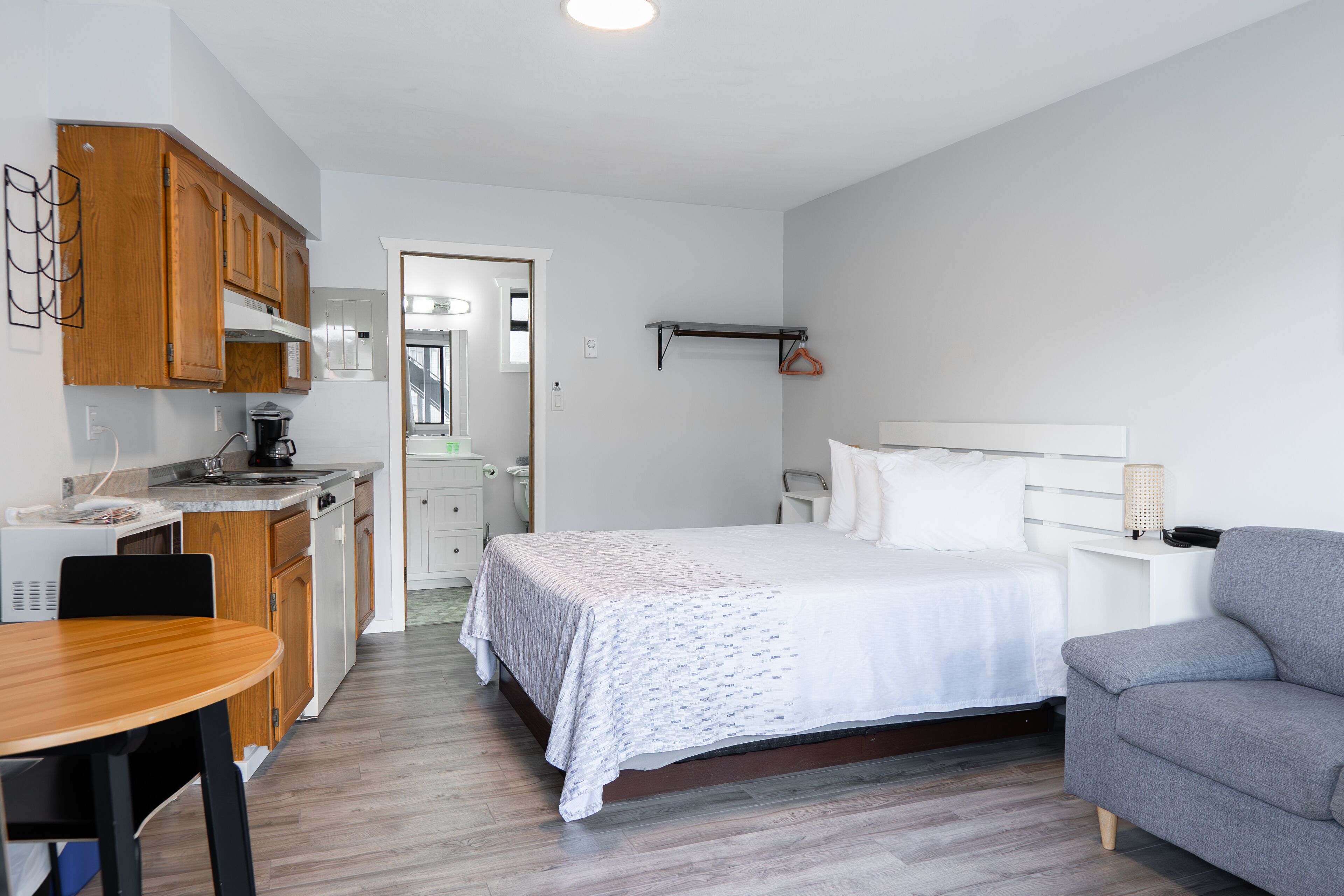 Una habitación de hotel pequeña con cama, un sofá gris, una mesa de comedor de madera y una kitchenette.