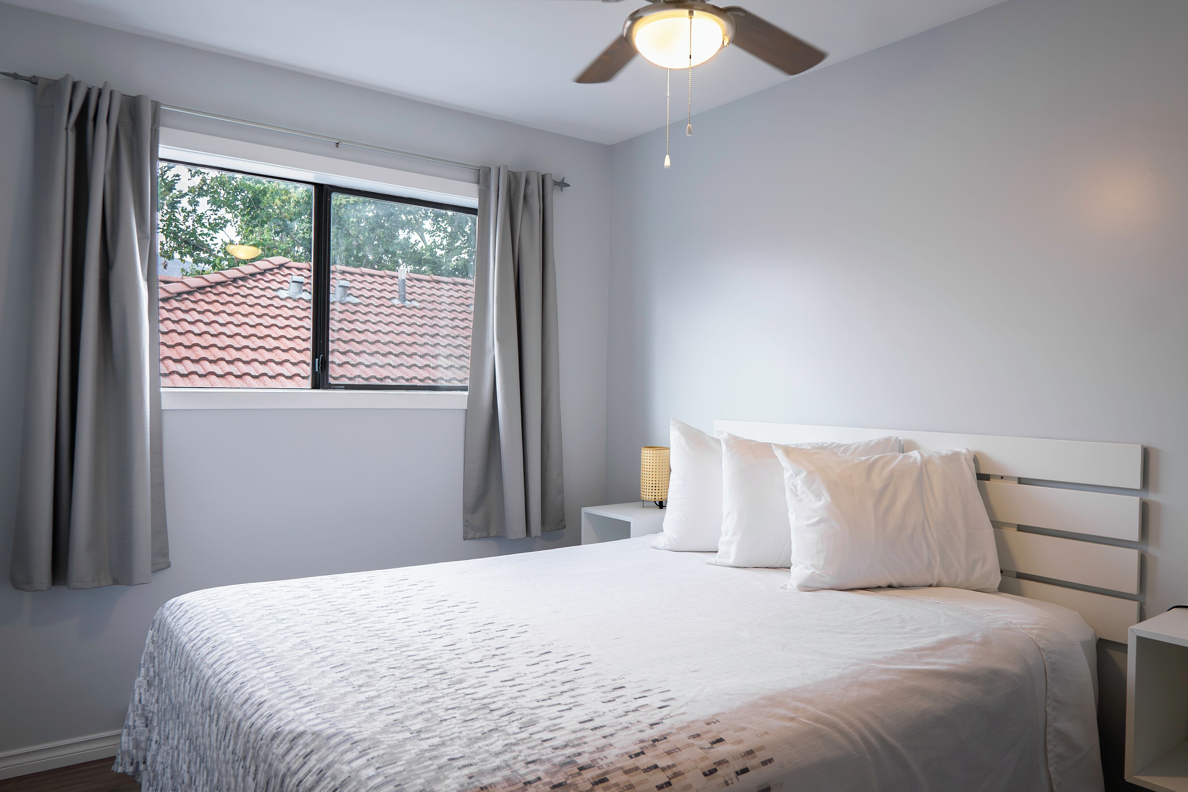A bedroom with a bed, a window with curtains, a nightstand with a lamp, and a ceiling fan.