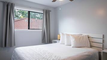 A bedroom with a bed, a window with curtains, a nightstand with a lamp, and a ceiling fan.
