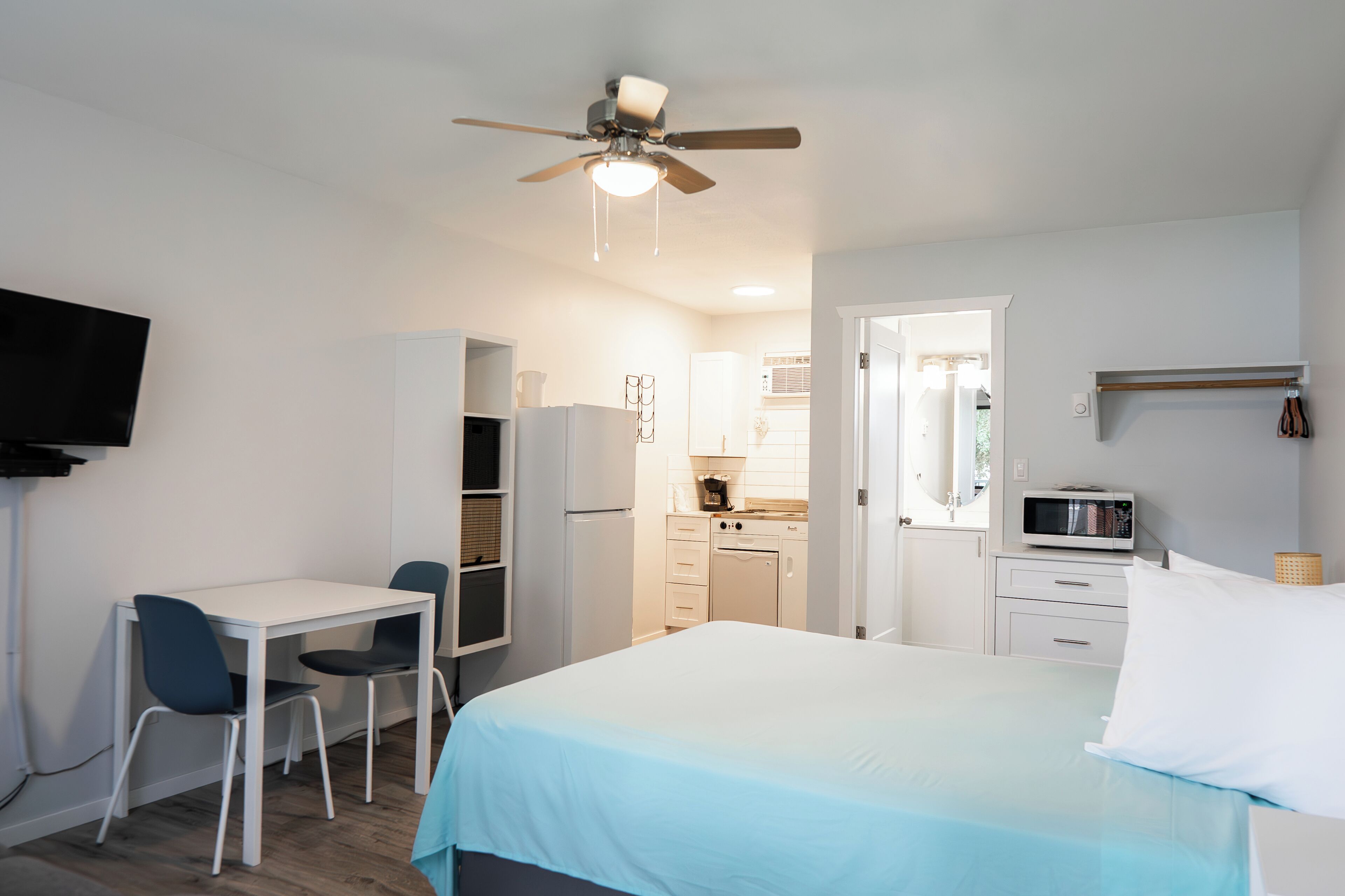 Una habitación de hotel compacta con cama, escritorio con sillas, kitchenette y ventilador de techo.