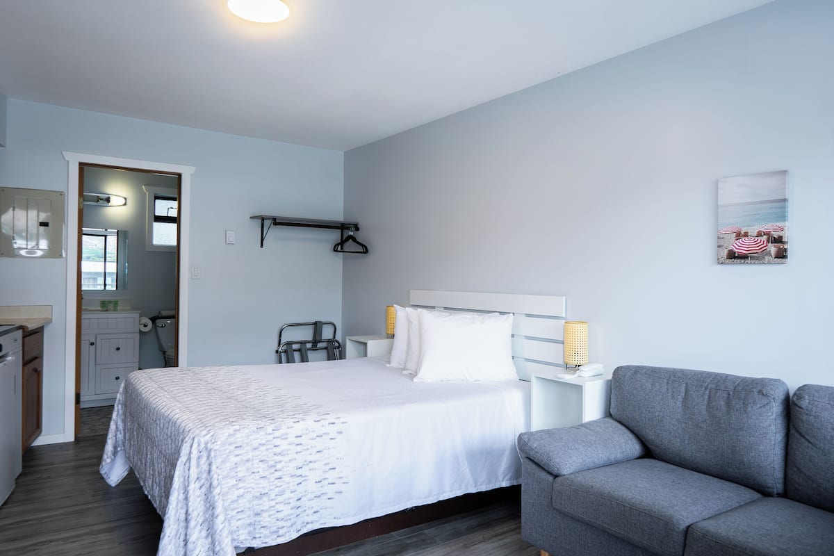 Un dormitorio con cama, sofá gris, mesita de noche con lámpara, estante en la pared y puerta que da a un baño.