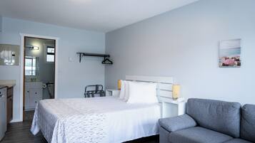 Un dormitorio con cama, sofá gris, mesita de noche con lámpara, estante en la pared y puerta que da a un baño.