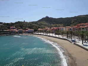 Uma vila costeira com praia, edifícios e uma colina com uma estrutura semelhante a um castelo.