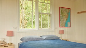 A bedroom with a large window, a bed with blue bedding, two bedside tables with lamps, and a map on the wall. - Romantic Cottage in the Blue Mountains with Valley Views (Hazelbrook)