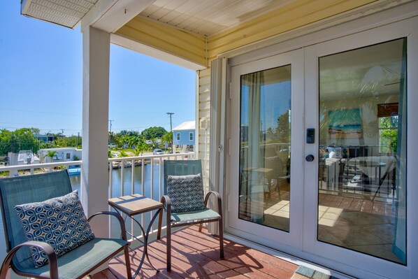 A covered outdoor area with two chairs and a table, a sliding glass door, and a view of the water and buildings in the background.