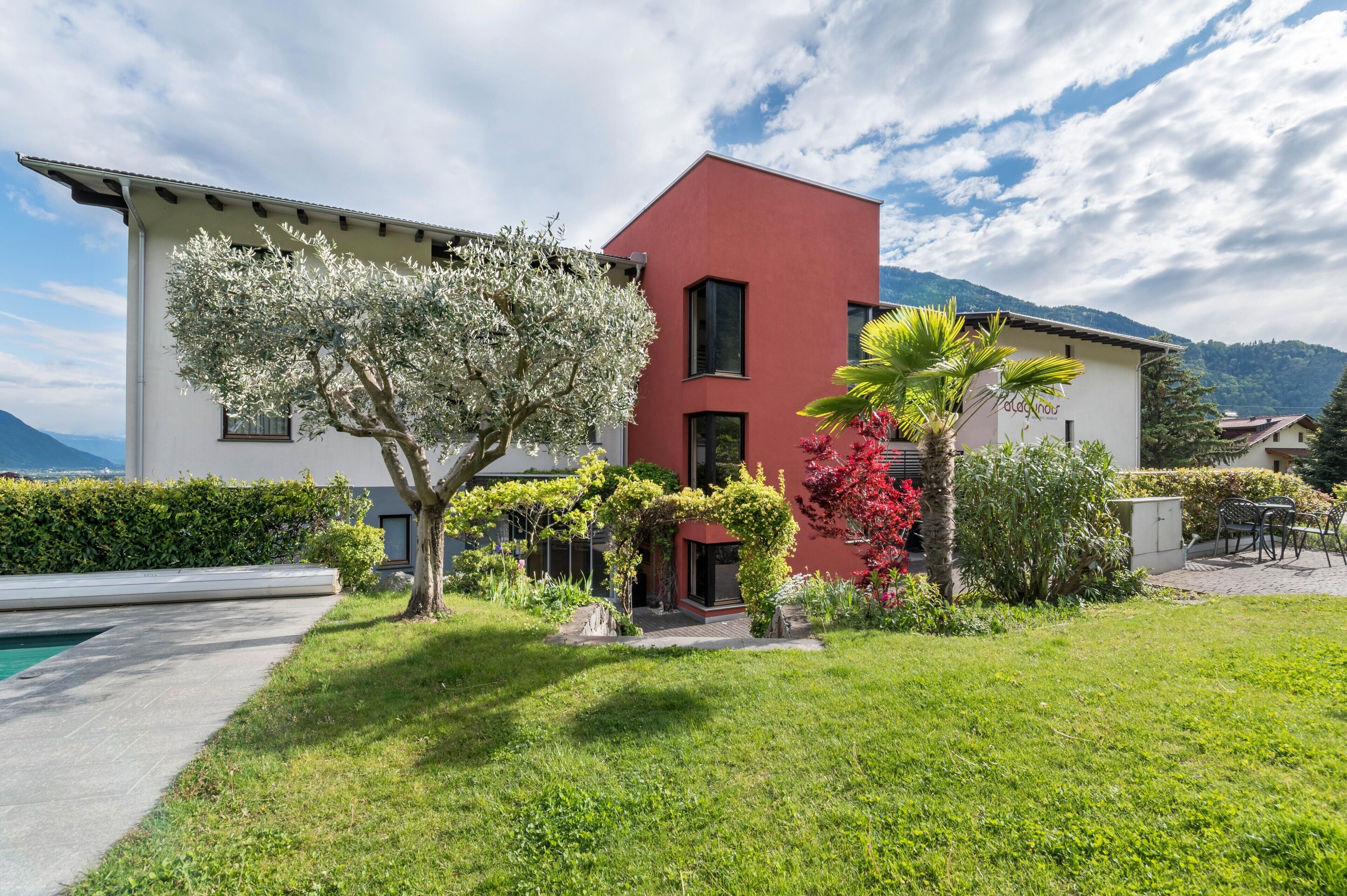 A modern two-story building with a red facade, a well-maintained garden with various plants and a small pool.