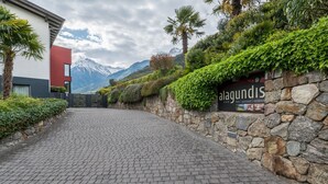 A paved pathway leading to a building with a stone wall and greenery, a sign reading 'alagundis,' and a mountain backdrop.