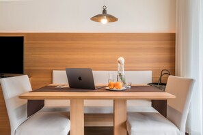 A modern dining area with a wooden table, white cushioned chairs, a laptop, a glass vase with flowers, and a fruit plate.