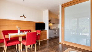 A modern kitchen with a flat-screen TV, wooden cabinets, and a dining area with red chairs and a white table. - Apartment 'Alagundis  Pyropus Familia' with Mountain View, Shared Pool and Wi-Fi (Lagundo)