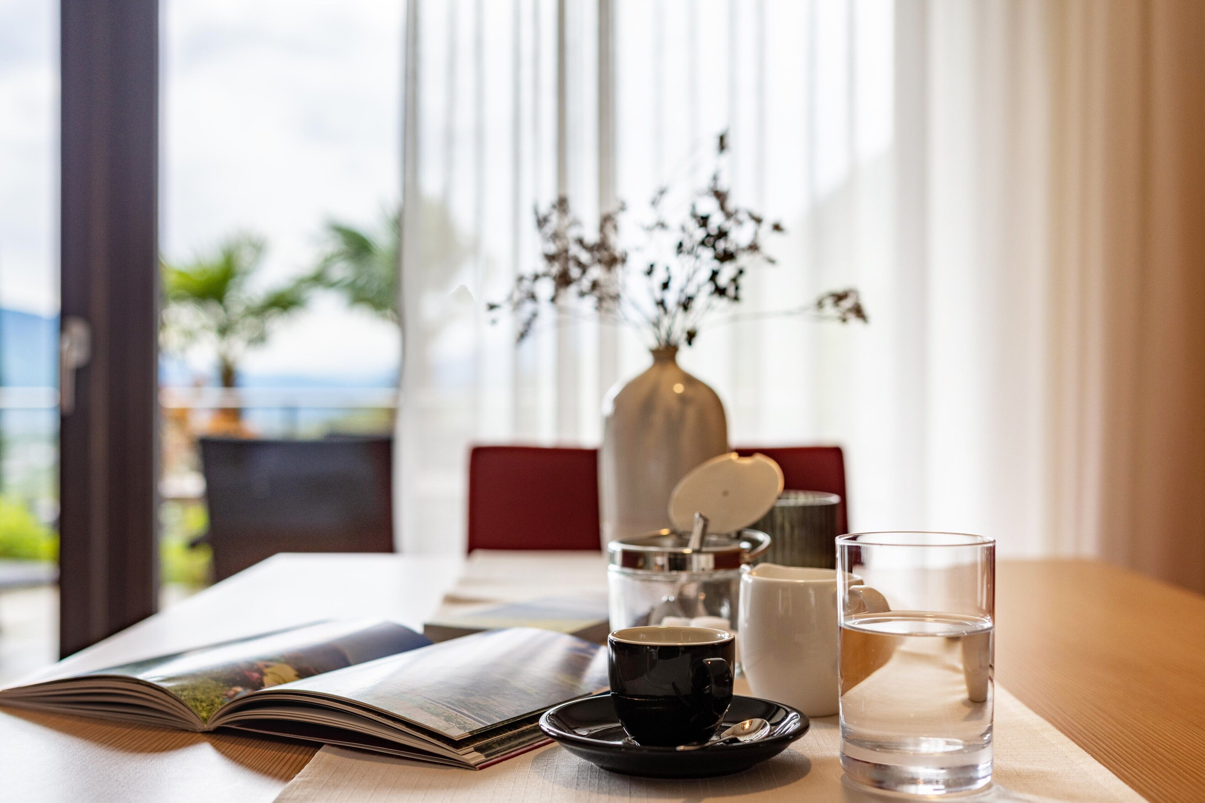 A table with a book, a cup, a glass of water, a vase with dried flowers, and a coffee maker.