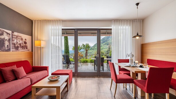 A modern hotel room with a red sofa, a dining table with chairs, a lamp, and a view of a mountainous landscape through the glass door. - Apartment 'Alagundis  Pyropus Familia' with Mountain View, Shared Pool and Wi-Fi (Lagundo)