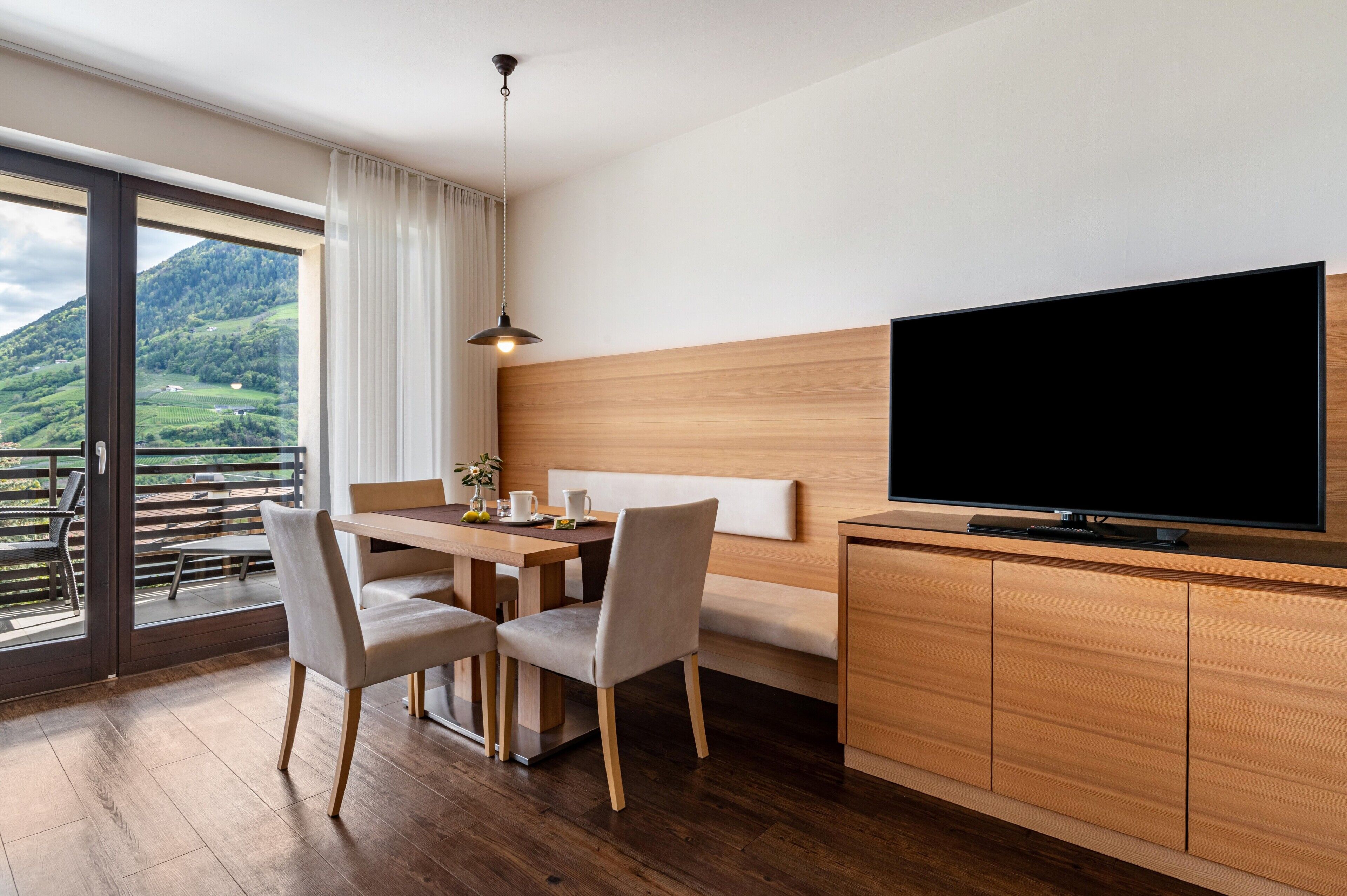 Ein modernes Zimmer mit einem Holztisch, zwei Stühlen, einem Fernseher und einem Balkon mit Blick auf grüne Hügel.