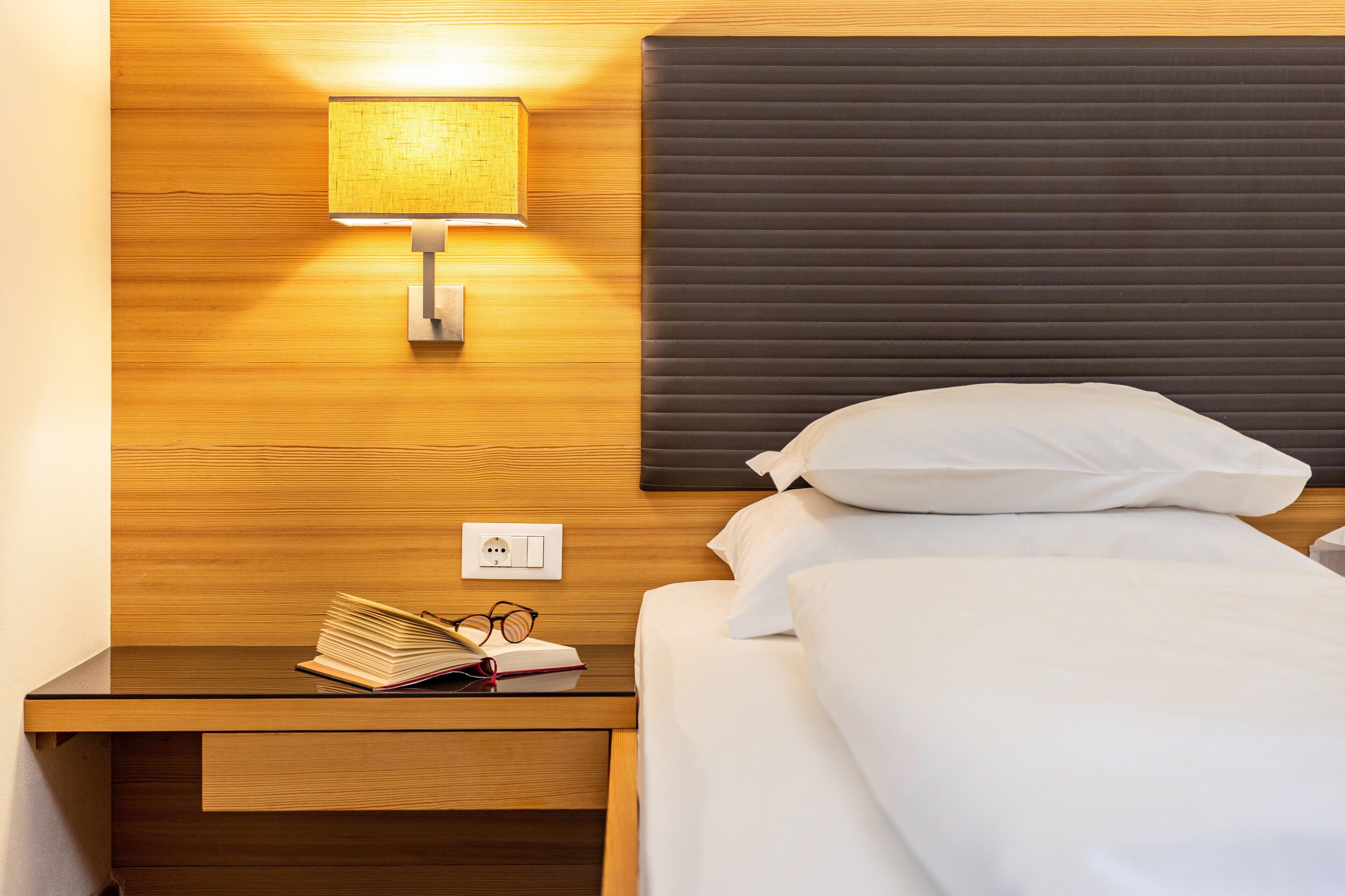 Hotelkamer met houten hoofdbord, een bedlamp, een bed met wit beddengoed, een nachtkastje met een boek en bril, en een stopcontact.