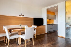Une cuisine moderne dotée d’un four encastré, un coin repas avec une table et des chaises en bois, et un téléviseur posé sur un meuble.