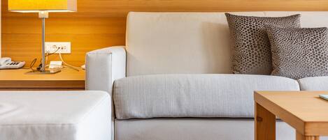 Una habitación de hotel moderna con cabecera de madera, sofá blanco, almohadas estampadas, mesita de noche con lámpara y una pequeña mesa de madera.