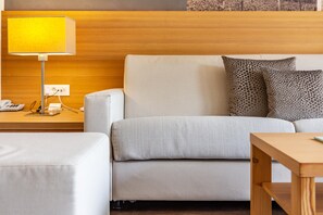 Une chambre d’hôtel moderne avec une tête de lit en bois, un canapé blanc, des coussins à motifs, une table de chevet en bois avec une lampe et une petite table en bois.