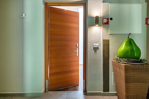 Un pasillo con una puerta de madera, una lámpara de pared, un detector de incendios y una planta verde en una cesta.