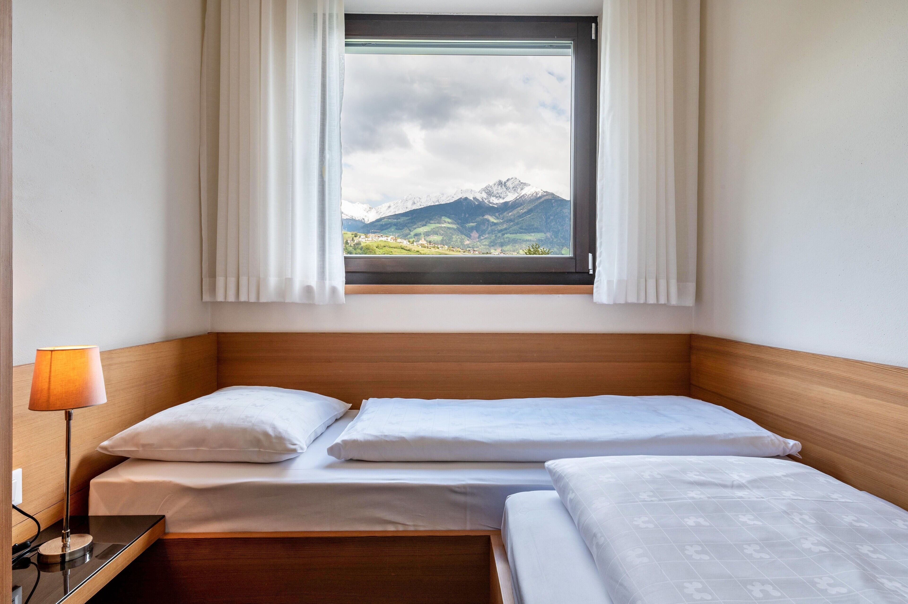 A hotel room with two beds, a window with curtains, a bedside lamp, and a view of mountains.