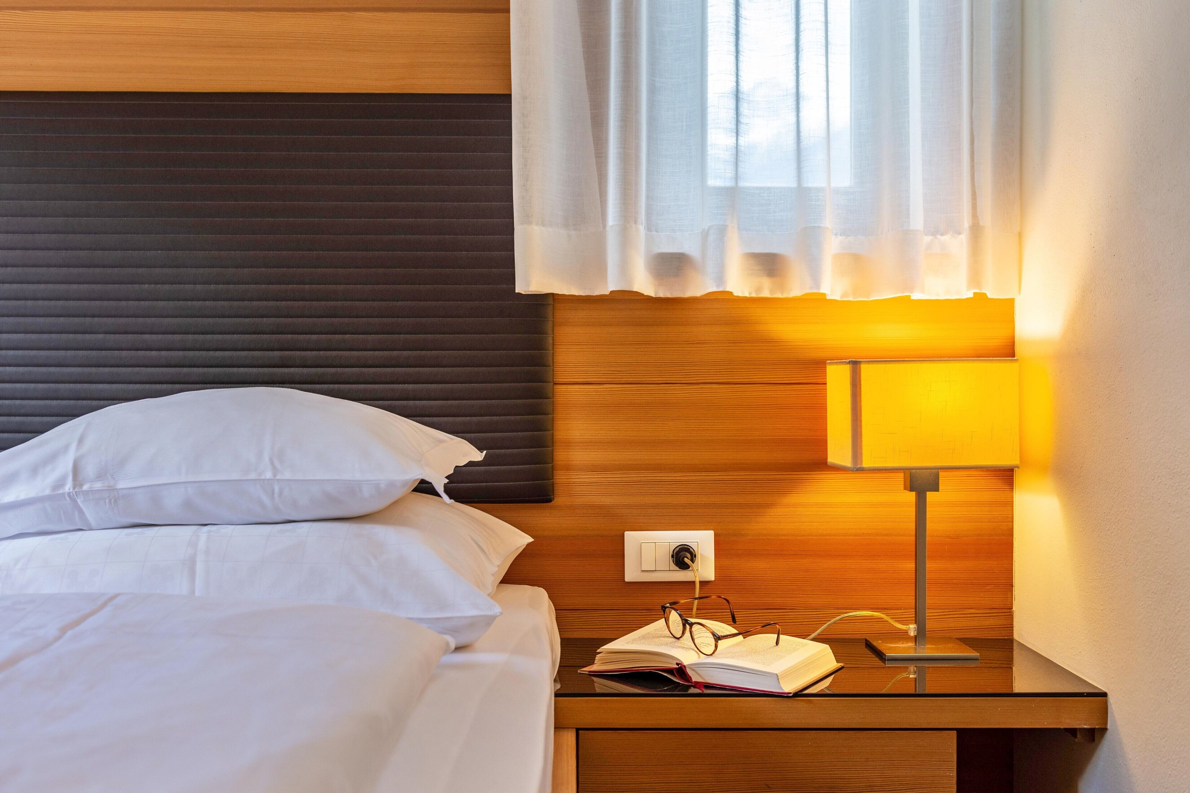 Una cama bien tendida con sábanas blancas, una mesita de noche con lámpara y una ventana con cortina blanca.