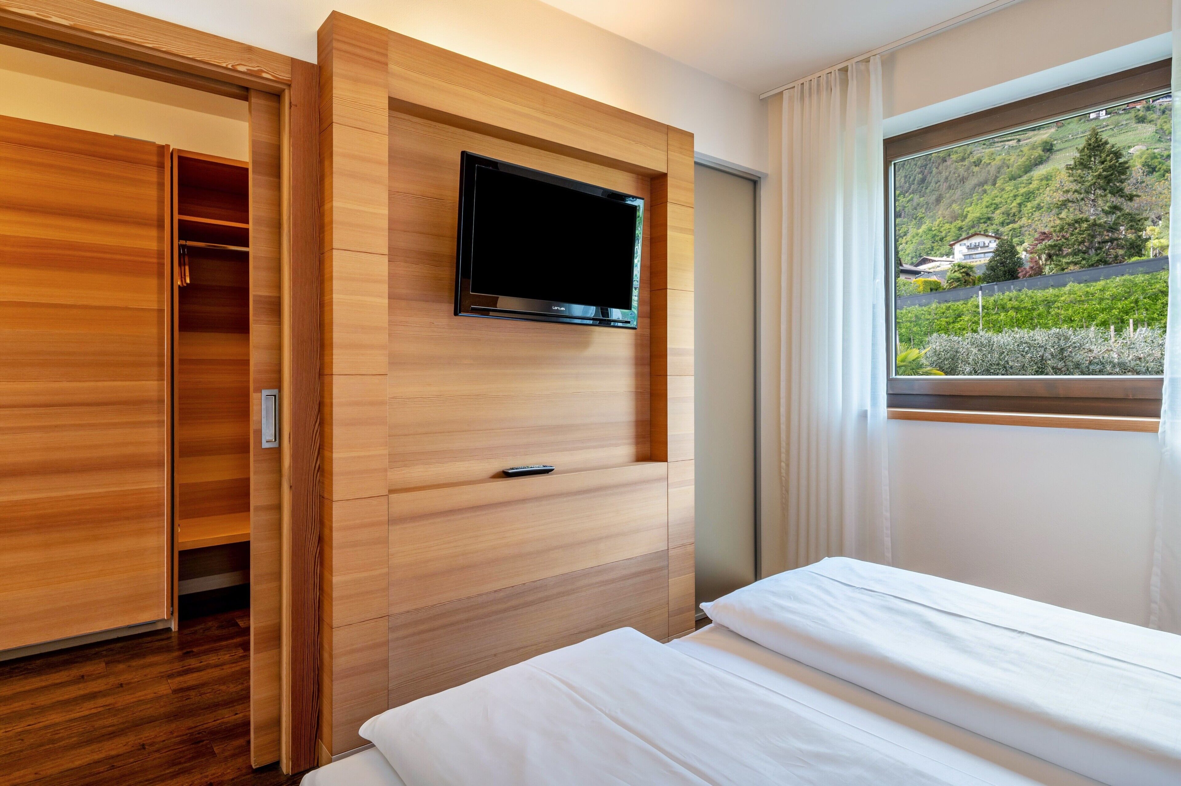 Quarto de hotel com cama, televisão na parede, guarda-roupa e janela com vista para vegetação e edifícios.