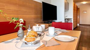 Quarto de hotel com área de jantar, mesa de madeira, tigela de frutas, vaso com flores e televisão de tela plana.