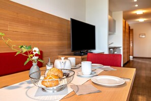 A hotel room with a dining area featuring a wooden table, a bowl of fruit, a vase with flowers, and a flat-screen TV.