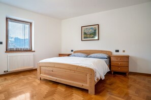 Une chambre à coucher avec un lit en bois, une fenêtre avec un rideau, un radiateur, une table de chevet, une commode et un tableau encadré au mur.