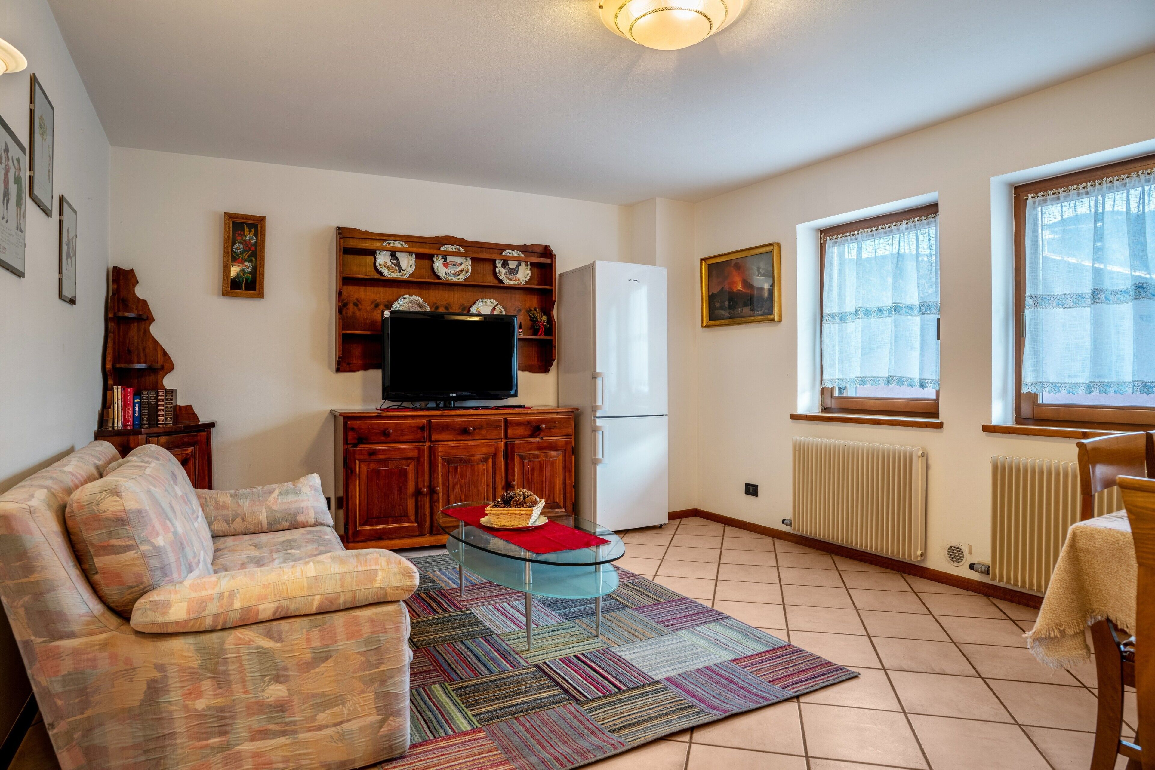 A living room with a sofa, a glass coffee table, a television, wooden cabinets, and a kitchen area with a refrigerator.