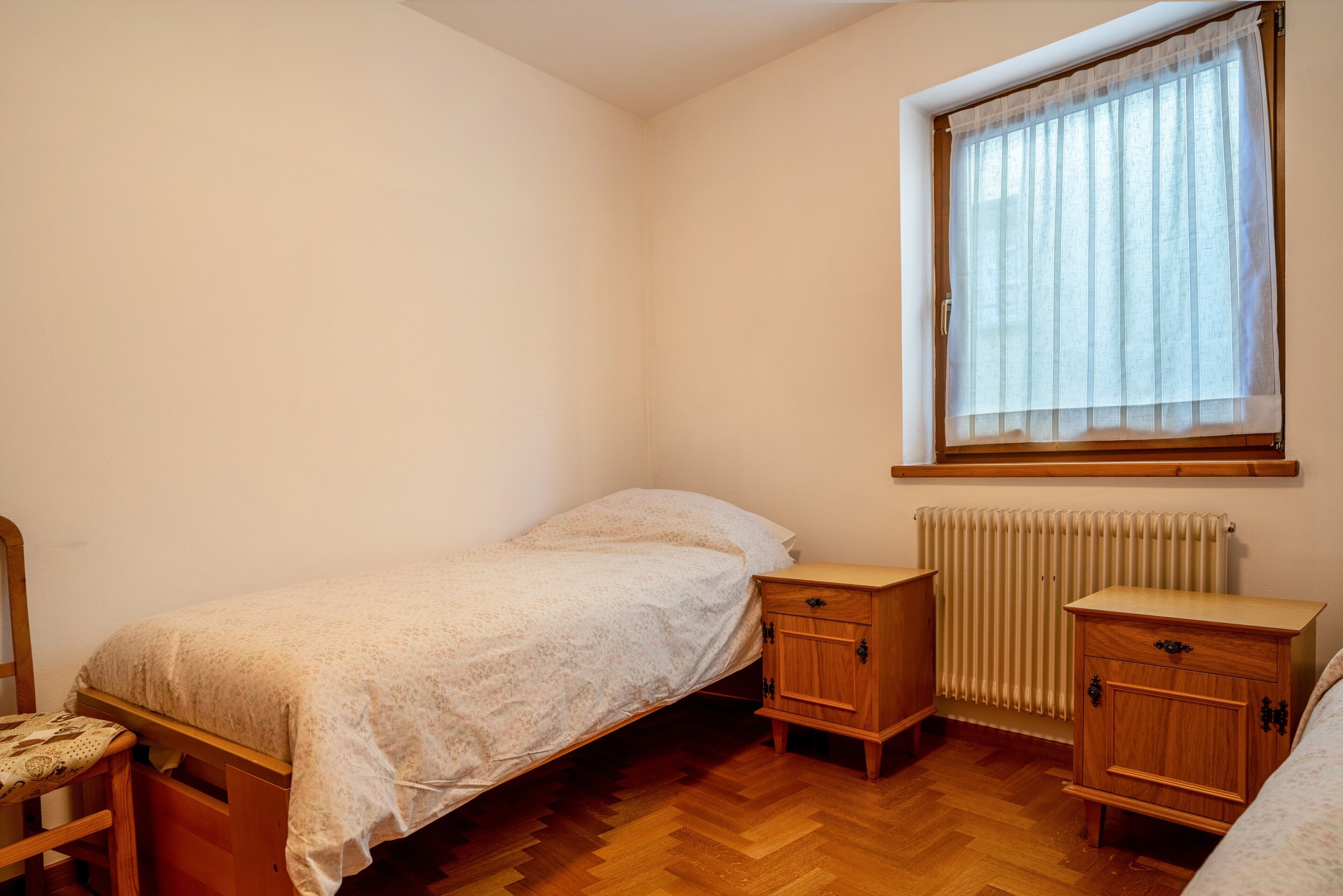 A small, single-bed room with a wooden floor, a radiator, a window with sheer curtains, and a wooden nightstand.