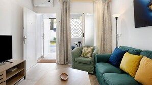 Ein Wohnzimmer mit Fernseher, einem grünen Sofa, einem Sessel mit Pflanzenkissen und einer Lampe.