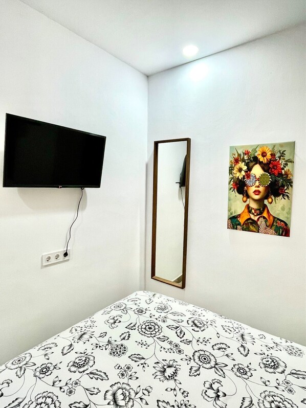 A room with a bed, a television mounted on the wall, a mirror, and a floral painting.