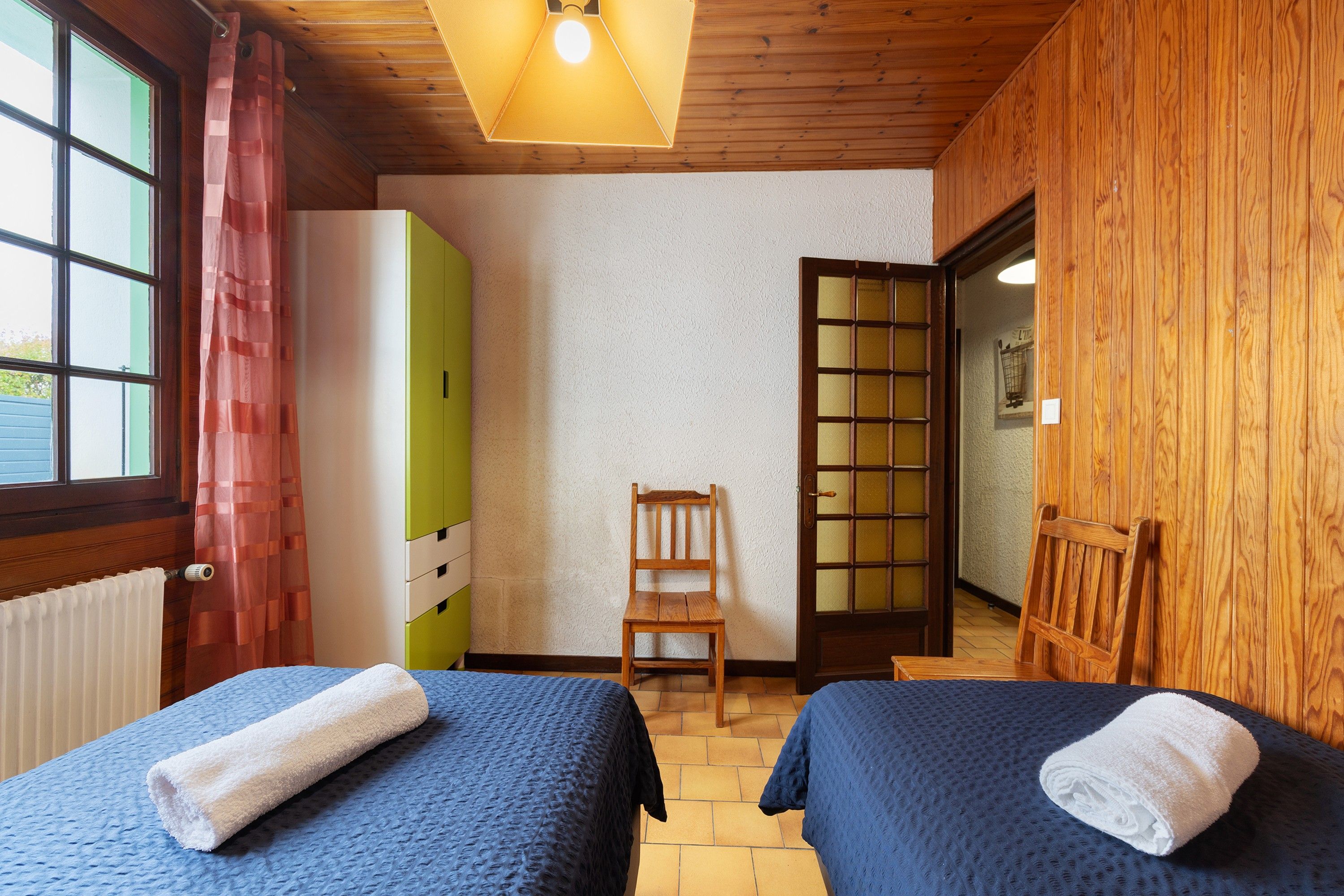 Quarto com duas camas, um guarda-roupa de madeira, uma cadeira e uma porta que dá para outro espaço.