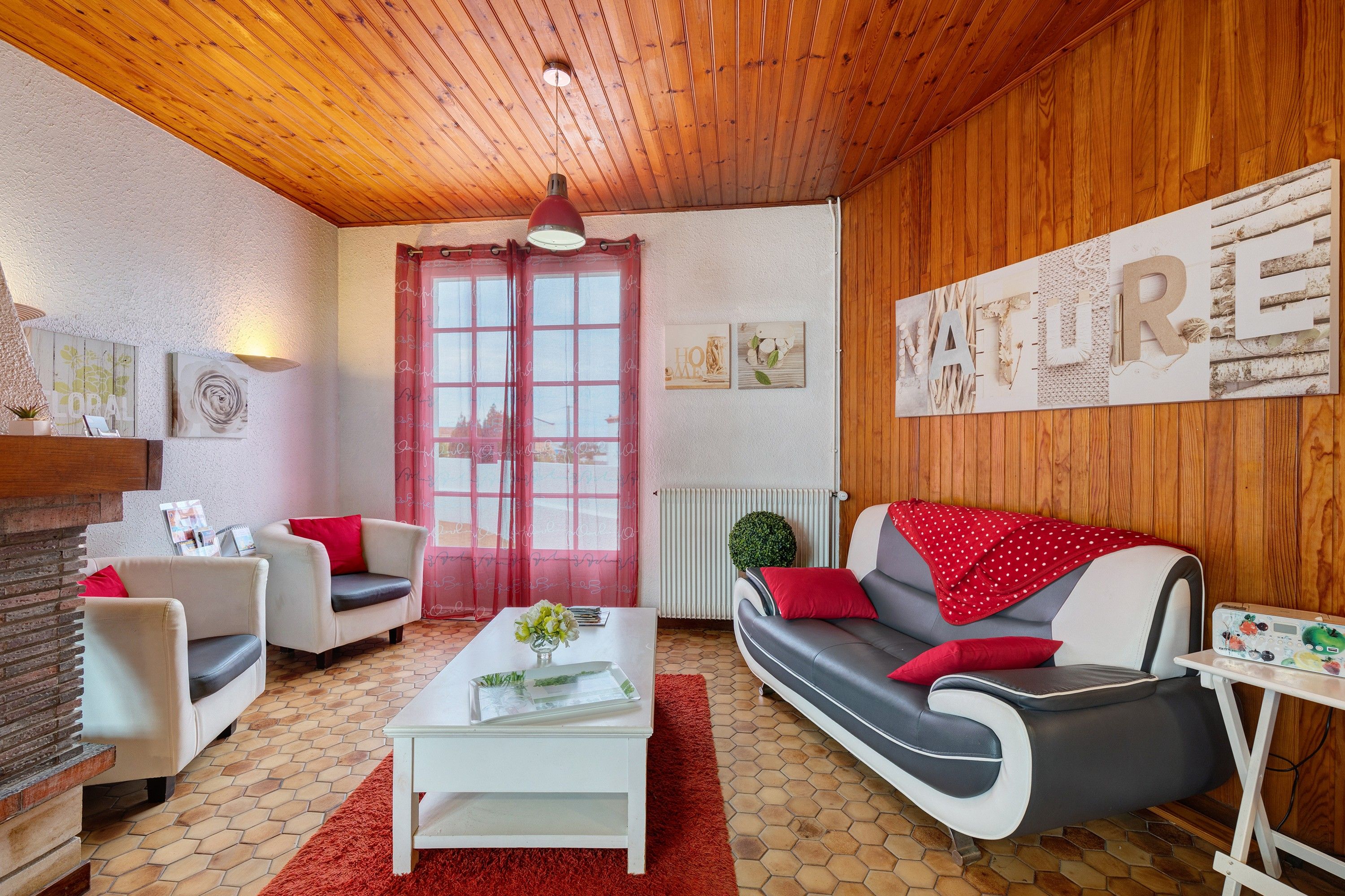Uma sala de estar aconchegante com teto de madeira, janela com moldura vermelha, sofá branco com almofadas vermelhas, mesa de centro branca e tapete vermelho.