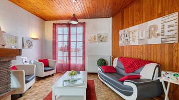 A cozy living room with a wooden ceiling, a red-framed window, a white sofa with red cushions, a white coffee table, and a red rug.