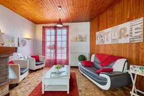 Een gezellige woonkamer met een houten plafond, een raam met rode omlijsting, een witte bank met rode kussens, een witte salontafel en een rood vloerkleed.
