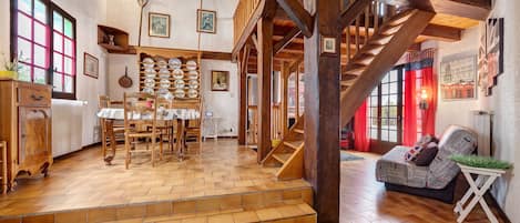 A rustic interior with wooden floors, a staircase, a dining area with a wooden table and chairs, and a living room with a sofa and a small table.