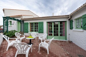 Une terrasse avec des chaises en plastique blanches et une petite table ronde, une porte verte et un bâtiment blanc avec des volets verts.