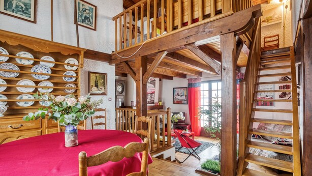 Een eetruimte met een rode tafelkleed, houten stoelen, een vaas met bloemen, en een trap die naar een entresol leidt.